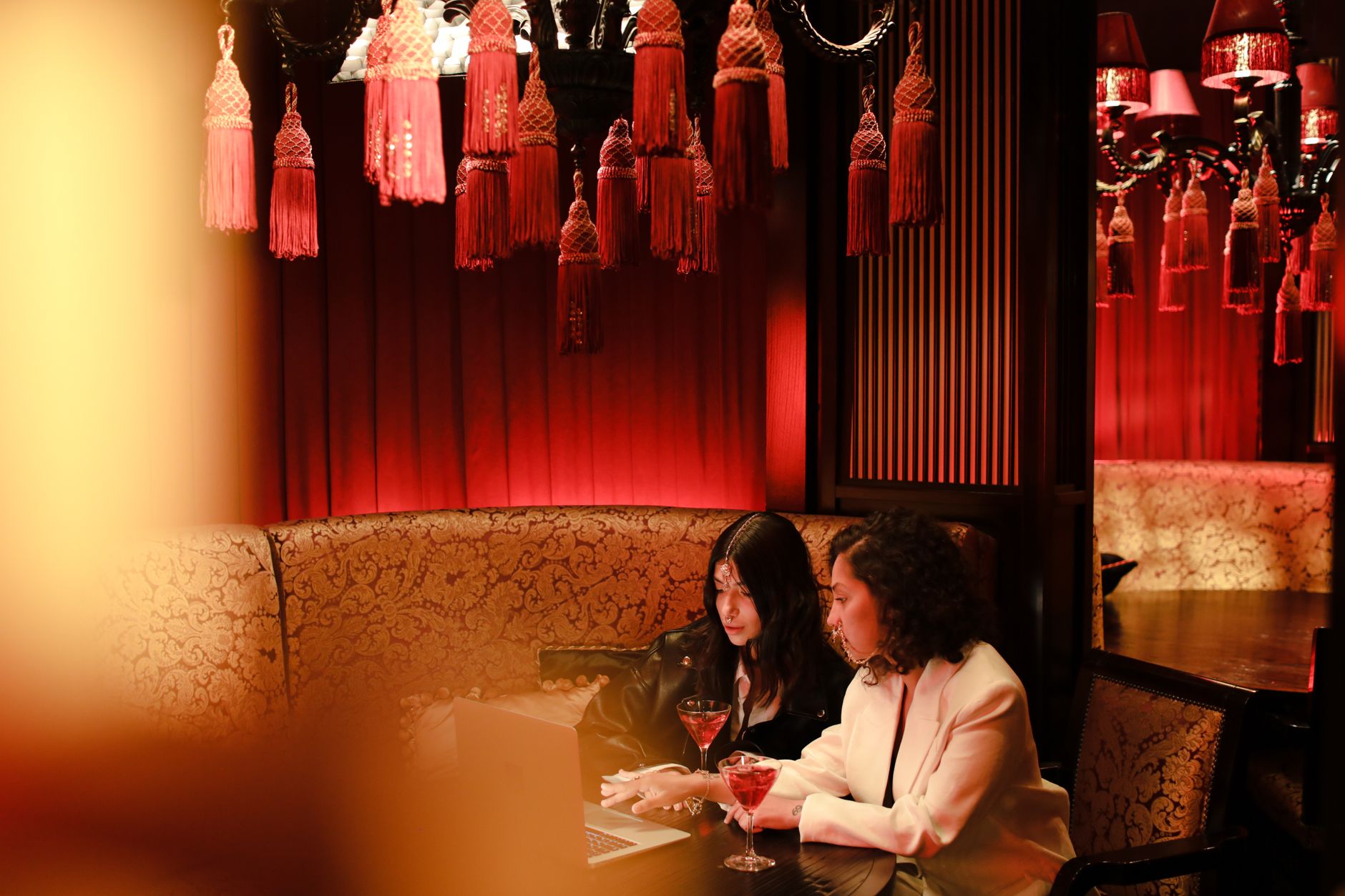 Two women working on a laptop over cocktails in a luxurious red-themed lounge.